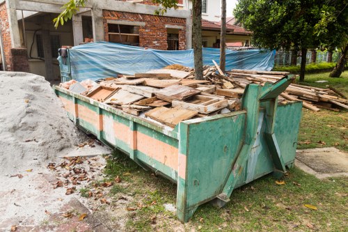A large green metal skip bin filled with a variety of flat, broken wooden planks and debris, situated on a patch of grass near a residential building under construction. The wooden planks are weathered, with some showing signs of paint or varnish, and are stacked haphazardly inside the bin. To the left of the skip, loose loose gravel and dirt are visible, while in the background, a blue construction tarp covers part of the building site, with exposed brickwork and unfinished walls. Adjacent to the skip are a few scattered leaves and small debris on the grass, indicating recent outdoor activity. The scene is outdoors, with trees and shrubbery in the background, and natural daylight illuminating the environment, emphasizing the clean but utilitarian nature of the rubbish removal process often handled by companies like Commercial Waste Hainault, serving local areas including the postcodes around the town.