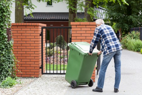 An older man with gray hair, dressed in a blue and white plaid shirt and blue jeans, is pushing a large green wheeled rubbish bin towards a gated driveway in a residential area. The gate is black metal and set between two brick pillars, leading to a garden and the front of a house. The driveway is made of light-colored paving stones, with some green plants and bushes along the edges. In the background, there are tall trees and a modern house exterior with a dark-colored roof and window frames. The scene appears to be during daytime with natural light, and the man seems to be preparing to dispose of waste as part of a rubbish collection service, demonstrating typical domestic rubbish removal activity near Hainault, Essex. Commercial Waste Hainault's services are implied by the context, focusing on waste collection for residential properties.