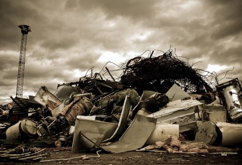 A large pile of discarded industrial and household waste materials is seen outdoors on a concrete or asphalt surface, with a cloudy sky overhead. The waste includes broken appliances such as washing machines and metal containers, along with twisted metal rods, plastic debris, and fragmented structural panels, all jumbled together in a haphazard manner. The pile extends from the foreground towards the background, where a tall metal tower and distant horizon are visible, giving an impression of an extensive waste disposal site. The materials exhibit a range of textures from smooth, painted surfaces to rough, corroded, and rusted metal and plastic fragments. The overall scene appears to be part of a rubbish clearance or waste collection site, possibly in an area servicing commercial waste removal in Hainault or nearby regions. The dark, stormy atmosphere underlines the industrial environment, emphasizing the importance of professional rubbish collection services provided by Commercial Waste Hainault. The image captures the cluttered, unorganized state of accumulated waste awaiting proper removal or disposal.